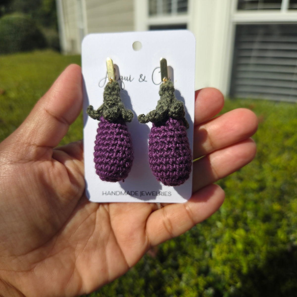 Handmade crochet purple and green earrings on a card held by a person outdoors.