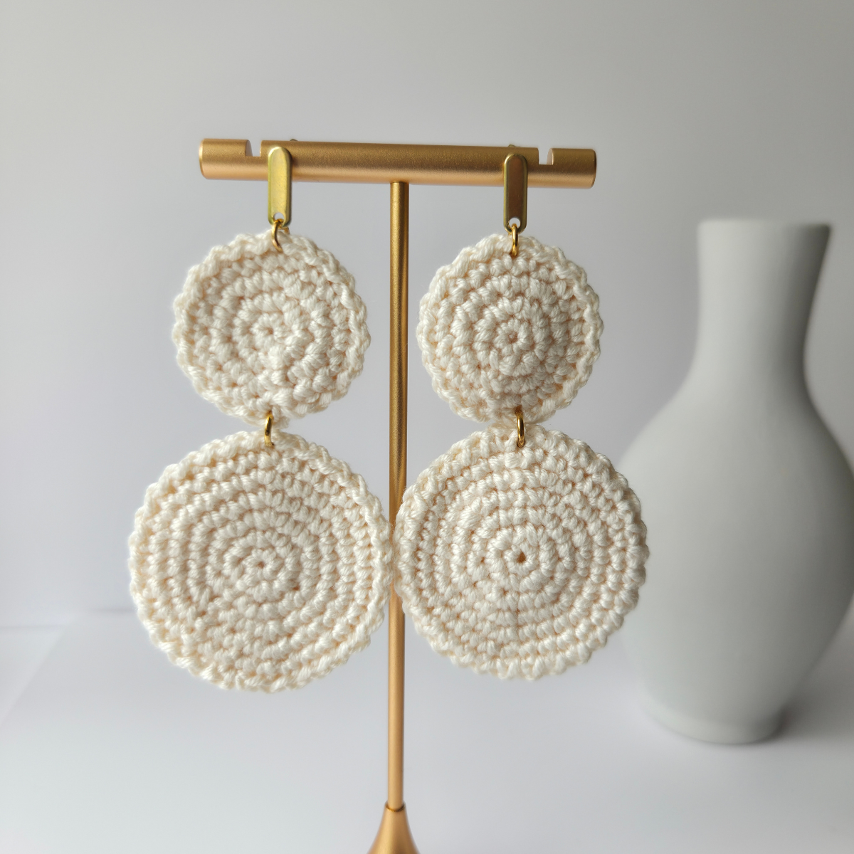 Cream-colored beaded earrings on a gold earring stand with a white vase in the background.