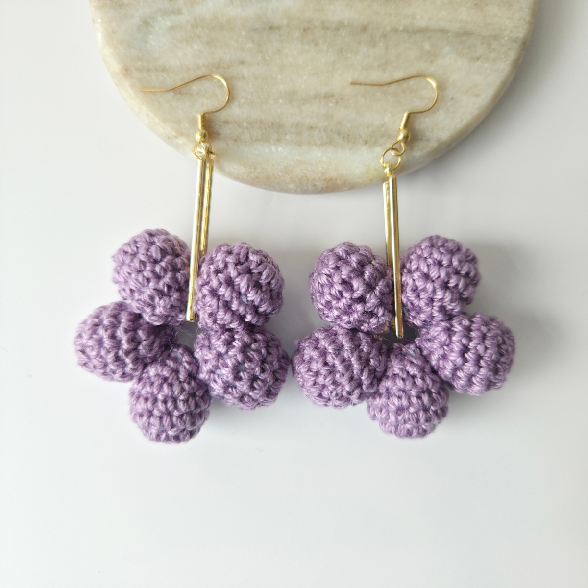 Purple crocheted earrings on gold hooks displayed on a white surface with a stone background.