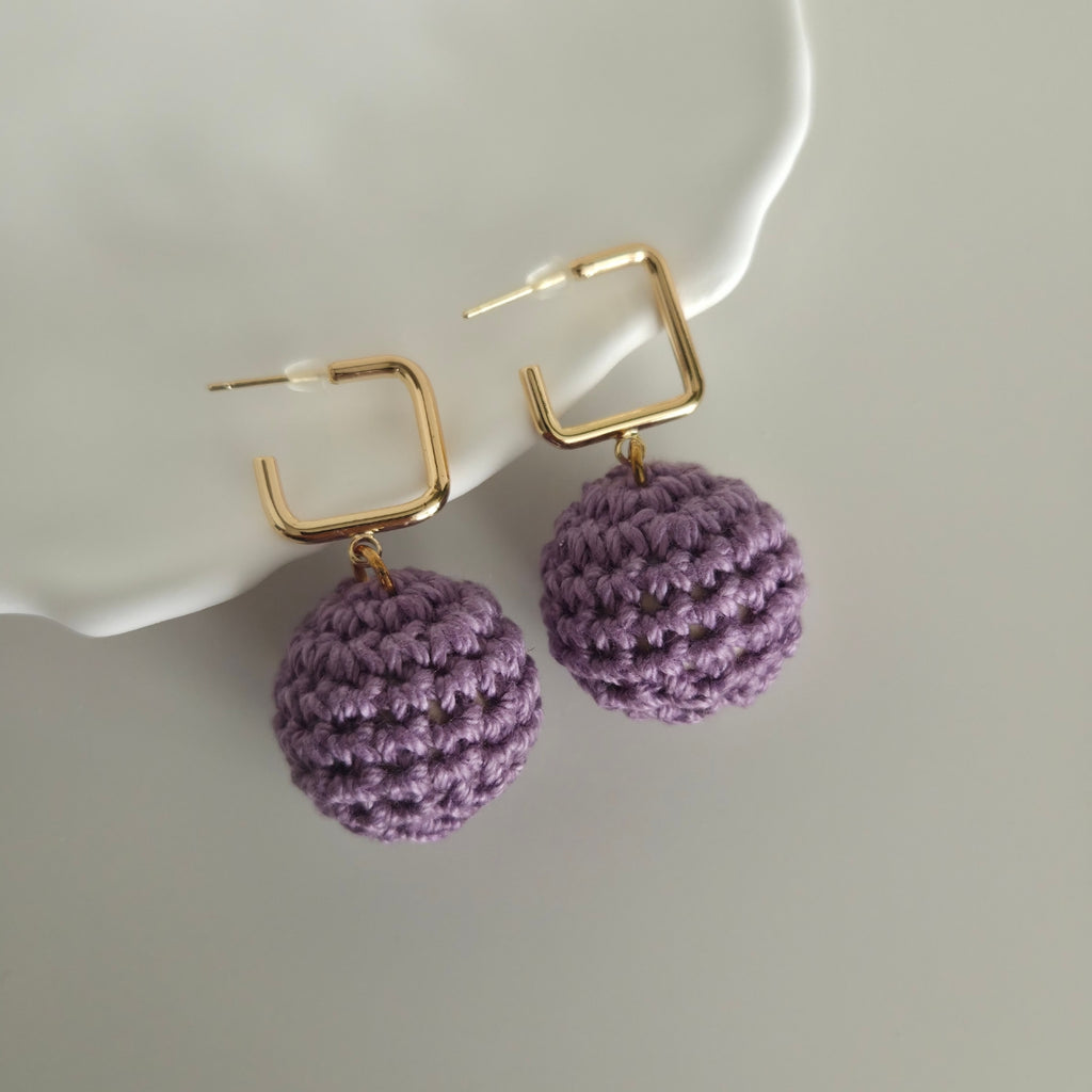 Purple crocheted ball earrings with gold square hoops on a white dish.