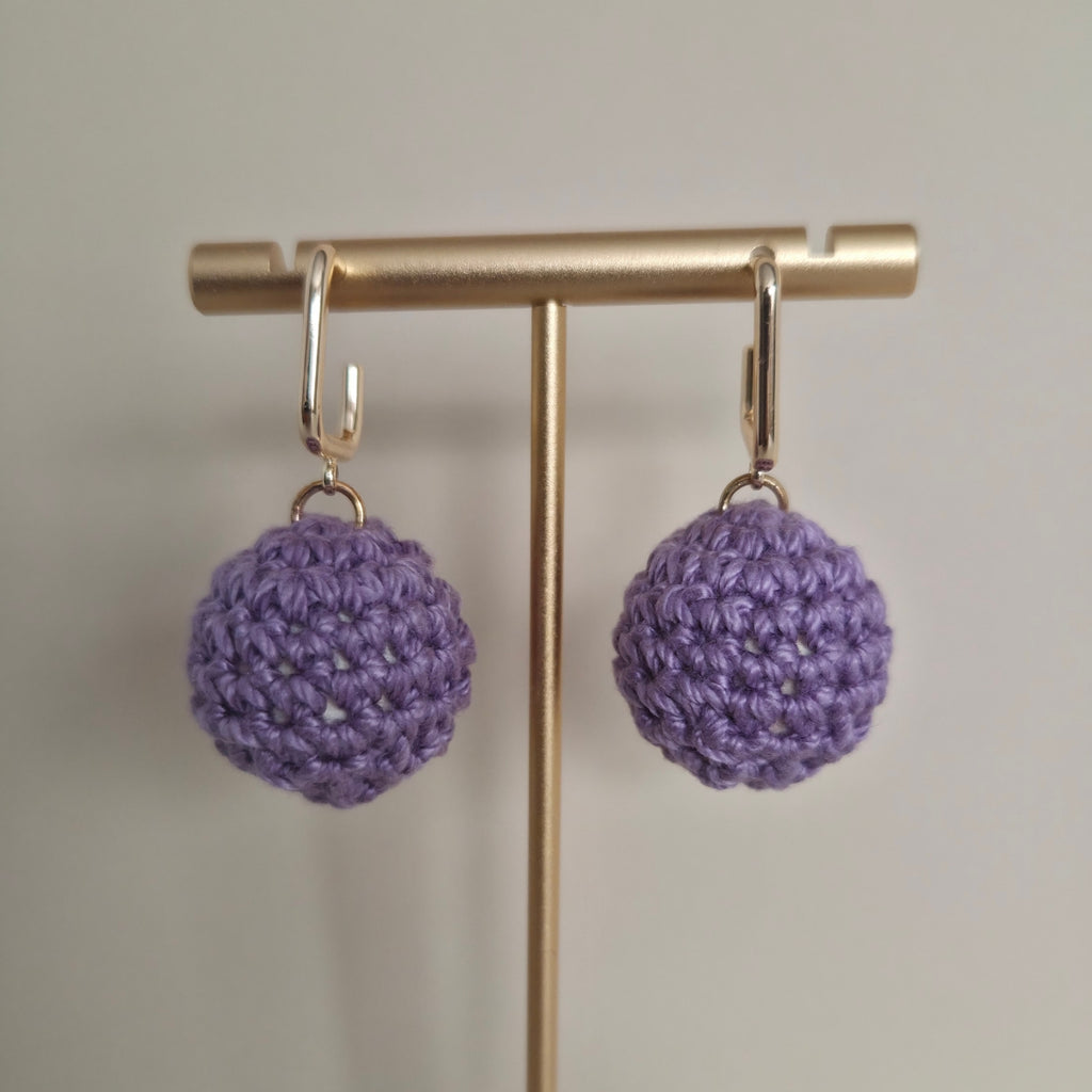 Purple crocheted earrings on a gold earring stand against a neutral background