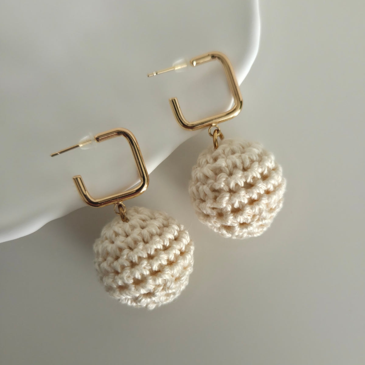Gold square hoop earrings with beige crocheted balls on a light gray background
