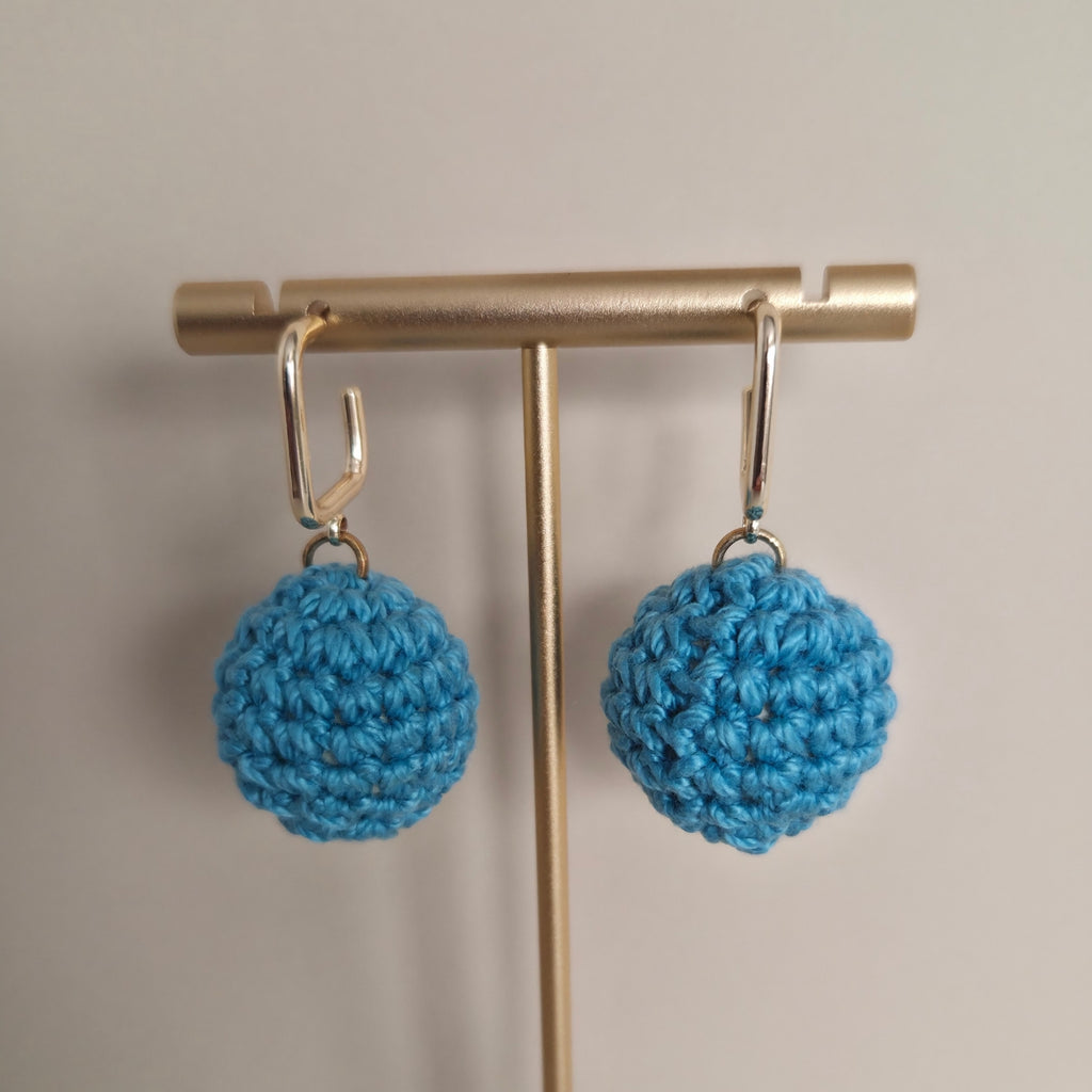 Blue crocheted ball earrings on a gold stand against a beige background