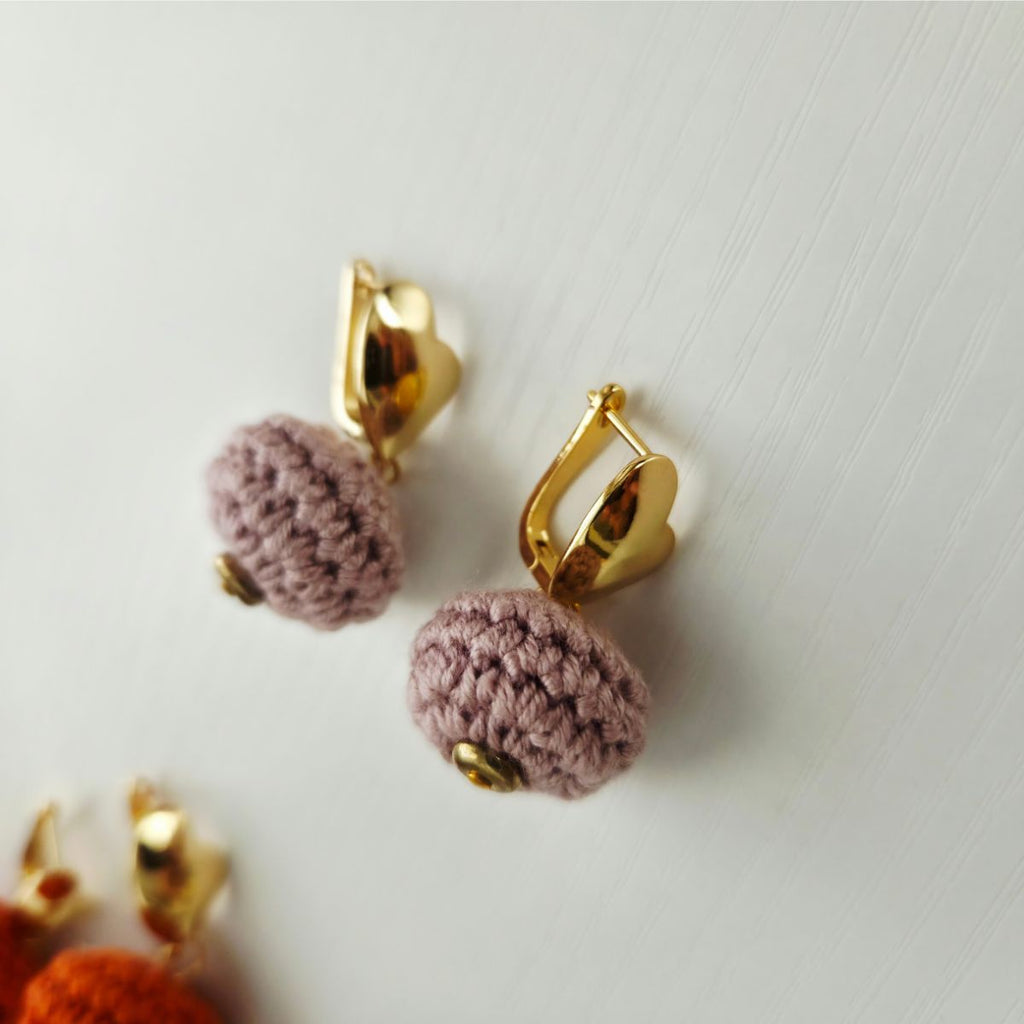 A pair of gold-toned huggie hoop earrings with small gold beads and dangling textured mauve-pink crochet pom-poms, photographed on a light neutral background.
