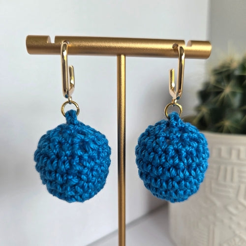 Blue handmade crochet ball drop earring on a gold earring stand with a blurred plant in the background.