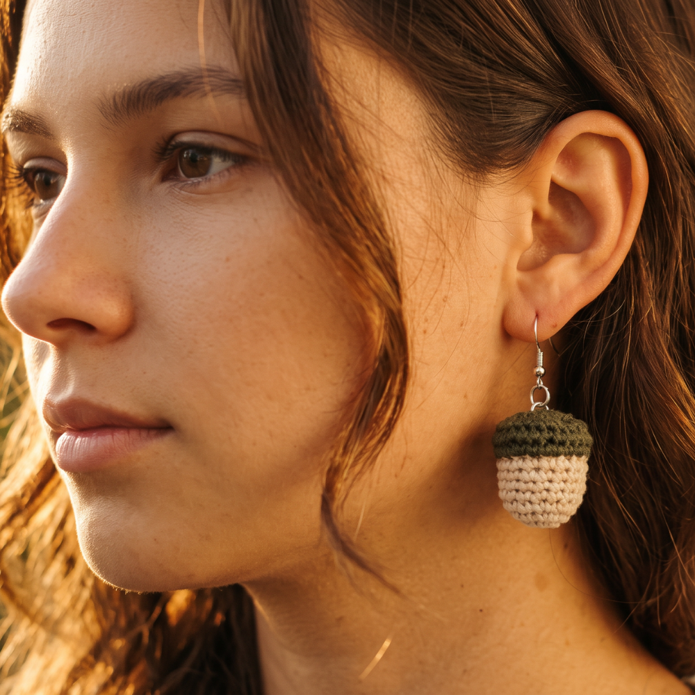 A close-up side profile of a woman wearing a handmade crochet acorn earring. The earring features a dark olive green top and a cream-colored bottom, suspended from a silver fishhook wire.