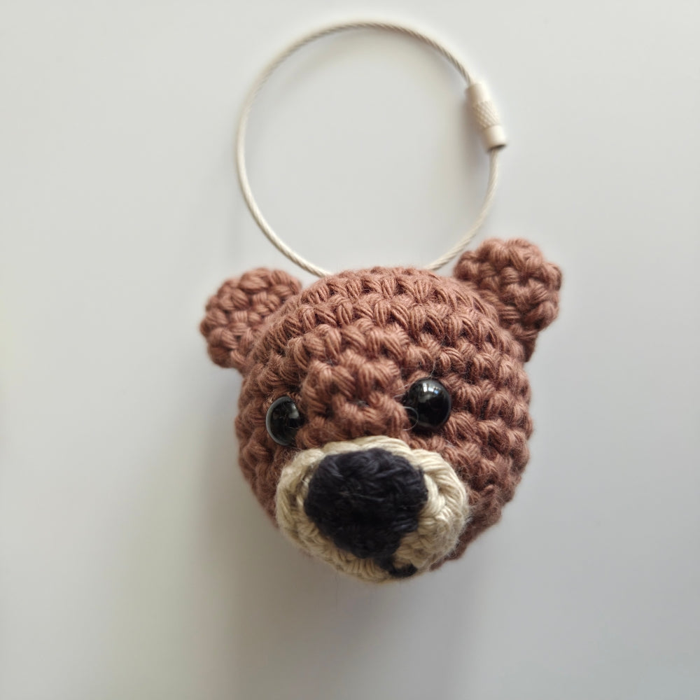 A crocheted bear head keychain bag in shades of brown, beige, and black, featuring a white plastic key ring attached at the top, photographed against a plain light gray background.