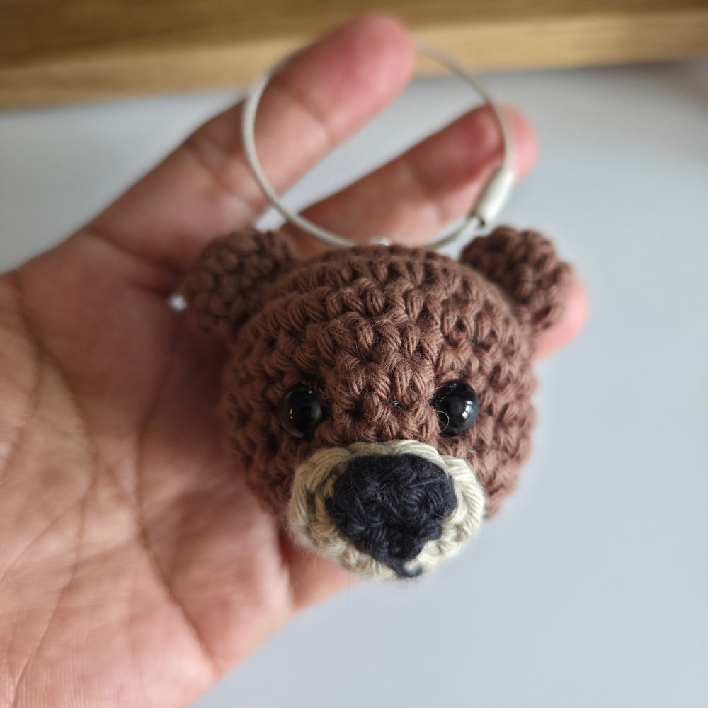A close-up of a hand holding a small crocheted bear head keychain or bag charm with a white metal ring. The bear is brown with black bead eyes, a cream-colored snout, and a black nose.