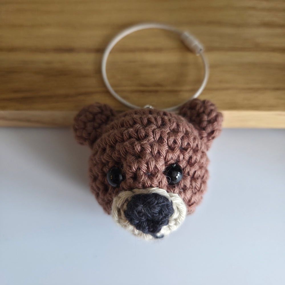 A handmade brown crocheted bear head keychain with black bead eyes, a black nose, and a cream-colored snout, attached to a white metal key ring, resting on a light gray surface with a wooden board in the background.
