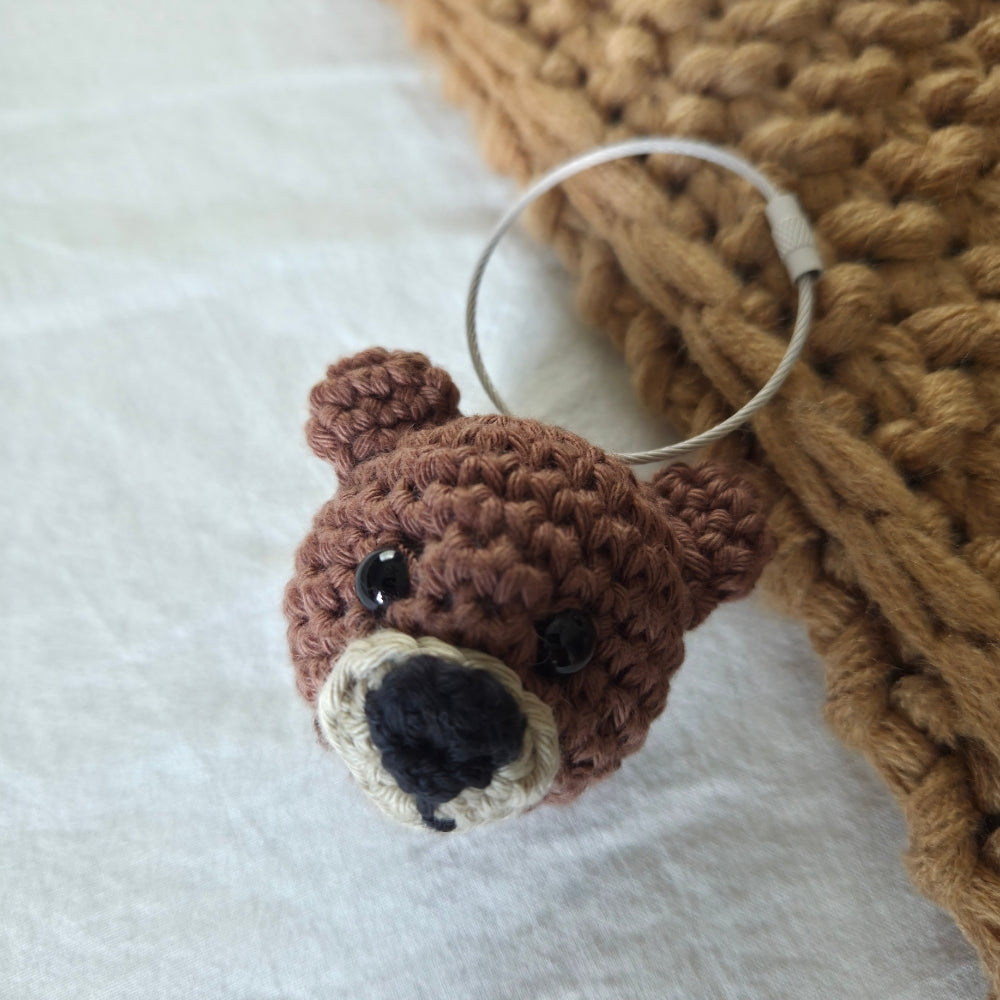 A small crocheted bear head keychain or bag charm with a silver metal ring, resting on a white fabric surface next to a chunky tan knitted blanket.