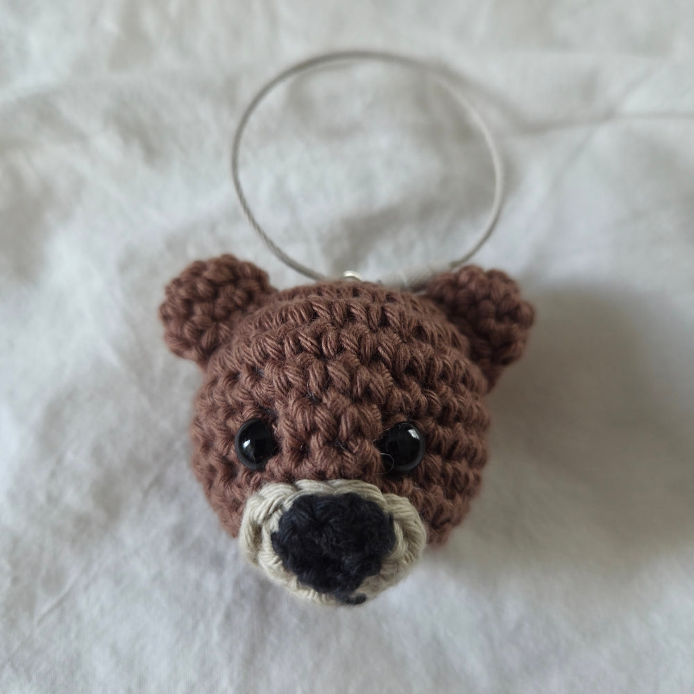 A small crocheted bear head keychain or bag charm with a clear plastic ring, featuring brown yarn, black bead eyes, and a cream and black snout, resting on a white fabric background.