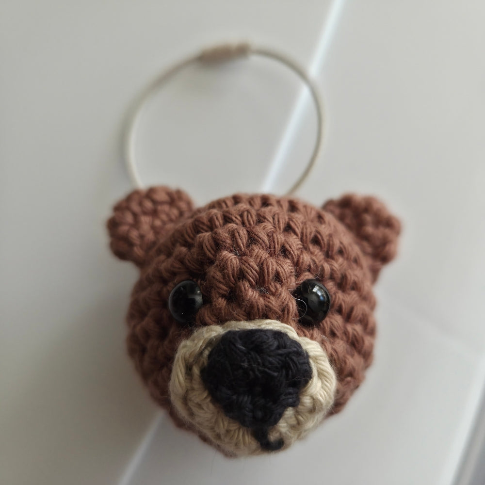 A brown crocheted bear head keychain with black bead eyes, a black nose, and a cream-colored snout, attached to a silver metal key ring.