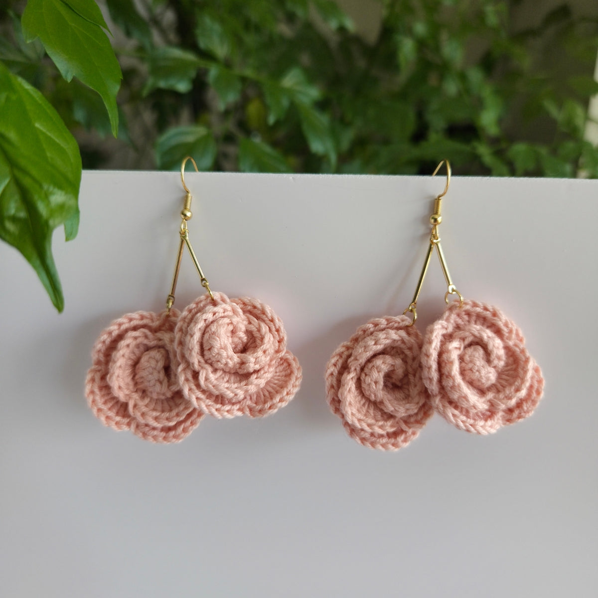 Pink crochet mini-rose dangle earrings on a white surface with green leaves in the background