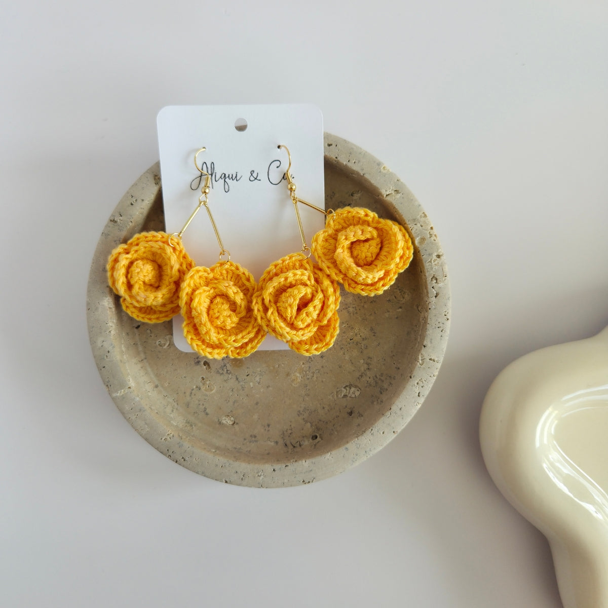 Pineapple yellow rose-shaped crochet earrings on a white card with 'Aliqui & Co' branding, displayed in a small concrete dish.