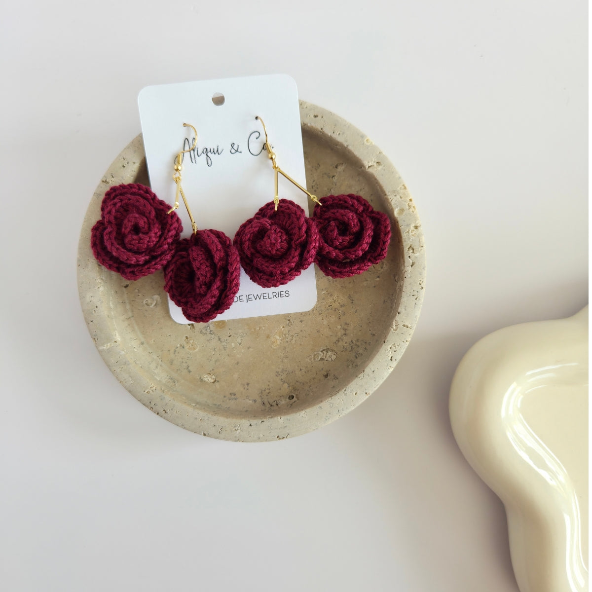 Wine red rose-shaped earrings on a white card with 'Aliqui & Co' branding, displayed in a beige dish.