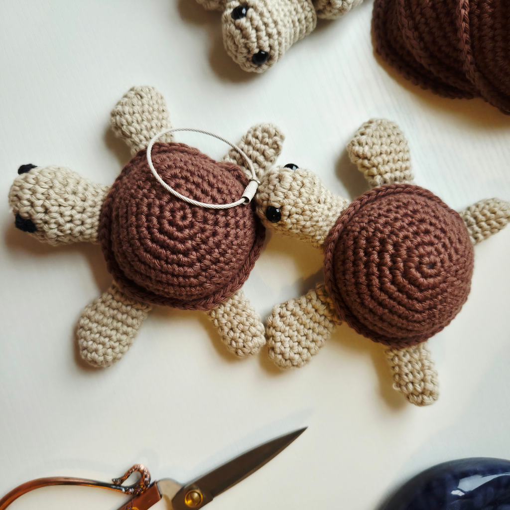 Two handmade crocheted turtle keychains on a white surface, one with a silver metal keyring. A pair of scissors is partially visible in the bottom left corner.