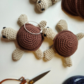 Two handmade crocheted turtle keychains on a white surface, one with a silver metal keyring. A pair of scissors is partially visible in the bottom left corner.