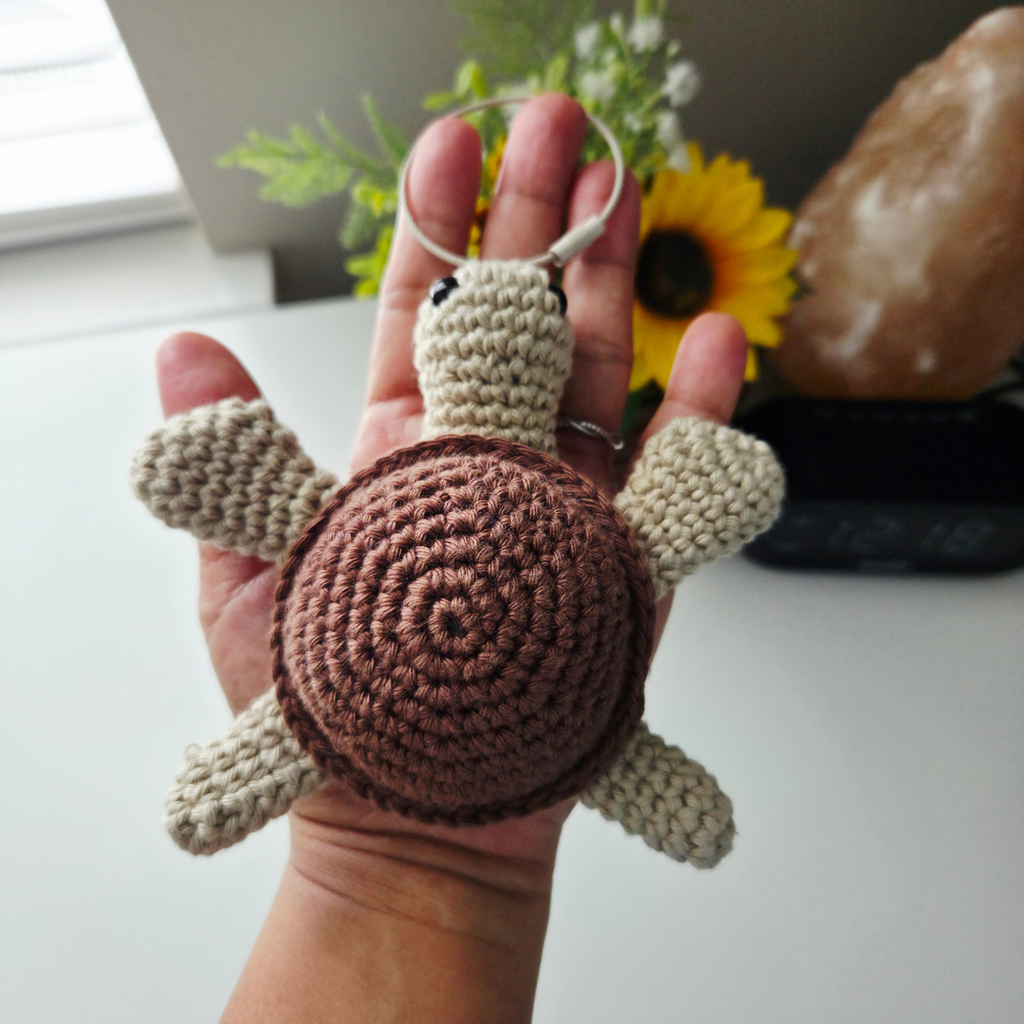 A handmade crocheted turtle keychain held in a person's hand. The turtle has a brown spiral shell and beige limbs and head, attached to a silver metal key ring.