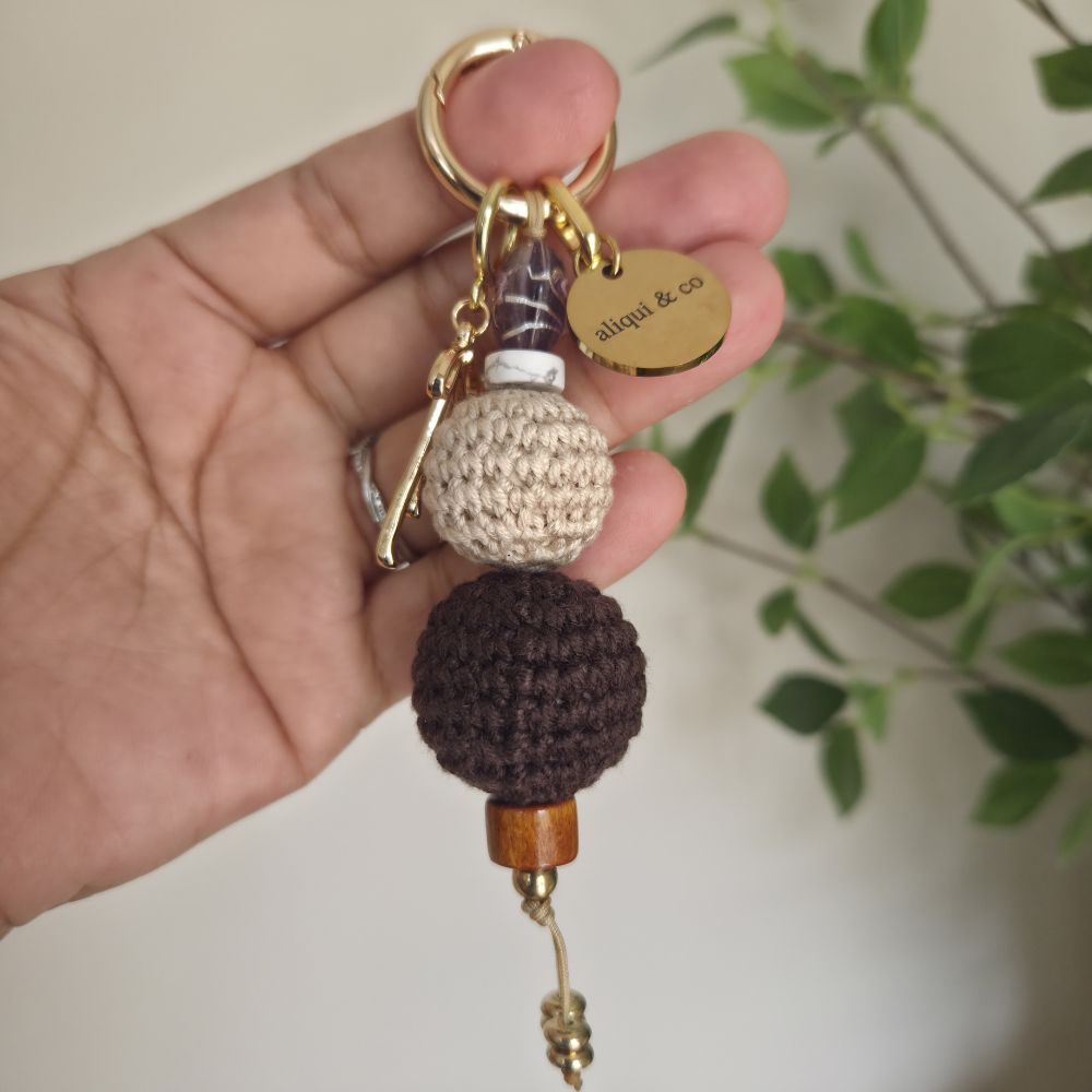 Hand holding a keychain with two crocheted balls and a gold tag, with a blurred plant in the background.