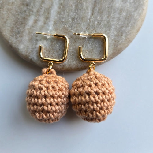 Pair of gold hoop earrings with light brown crocheted balls on a stone surface.