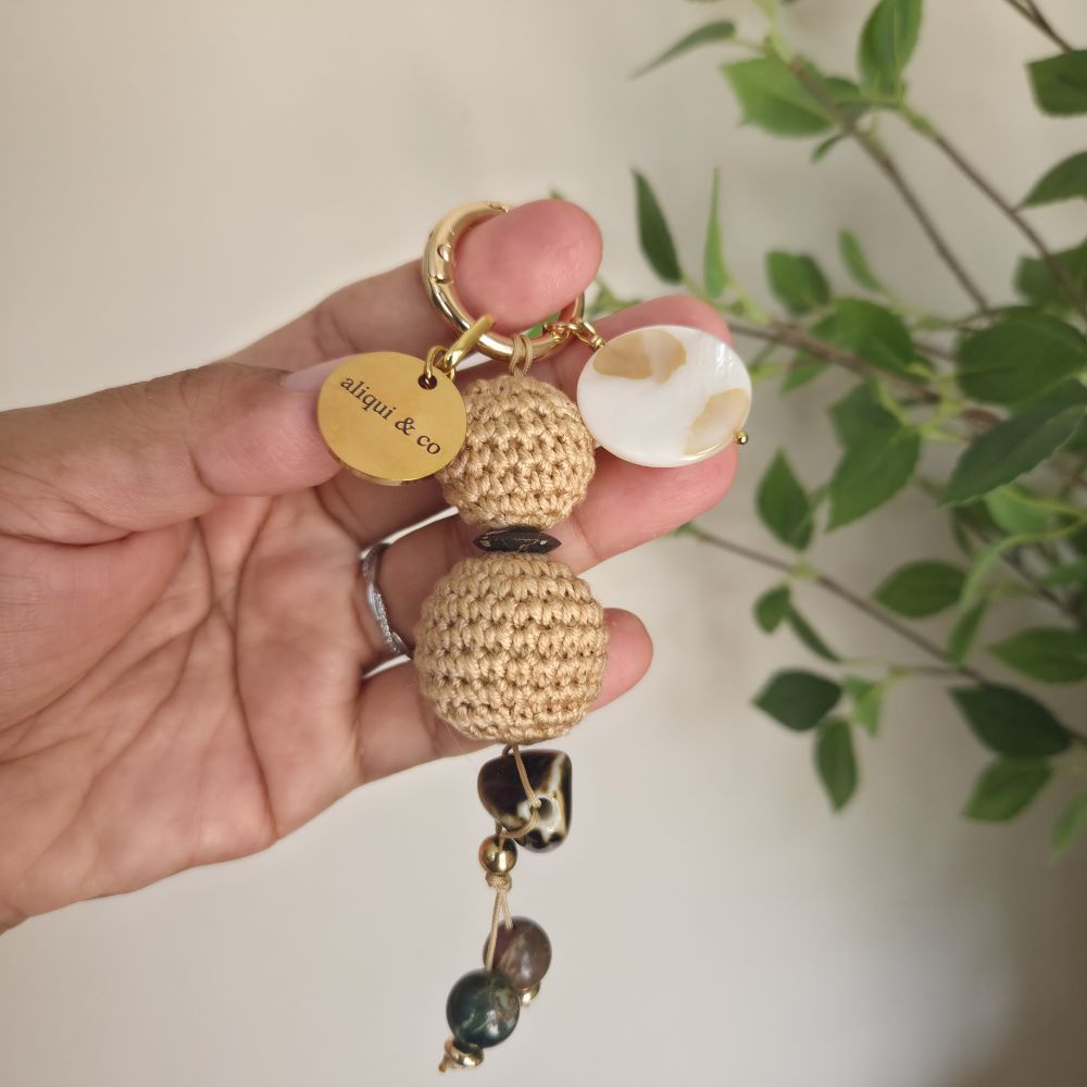 Hand holding a keychain with spherical beads and a 'aliqui & co' tag against a blurred green plant background.