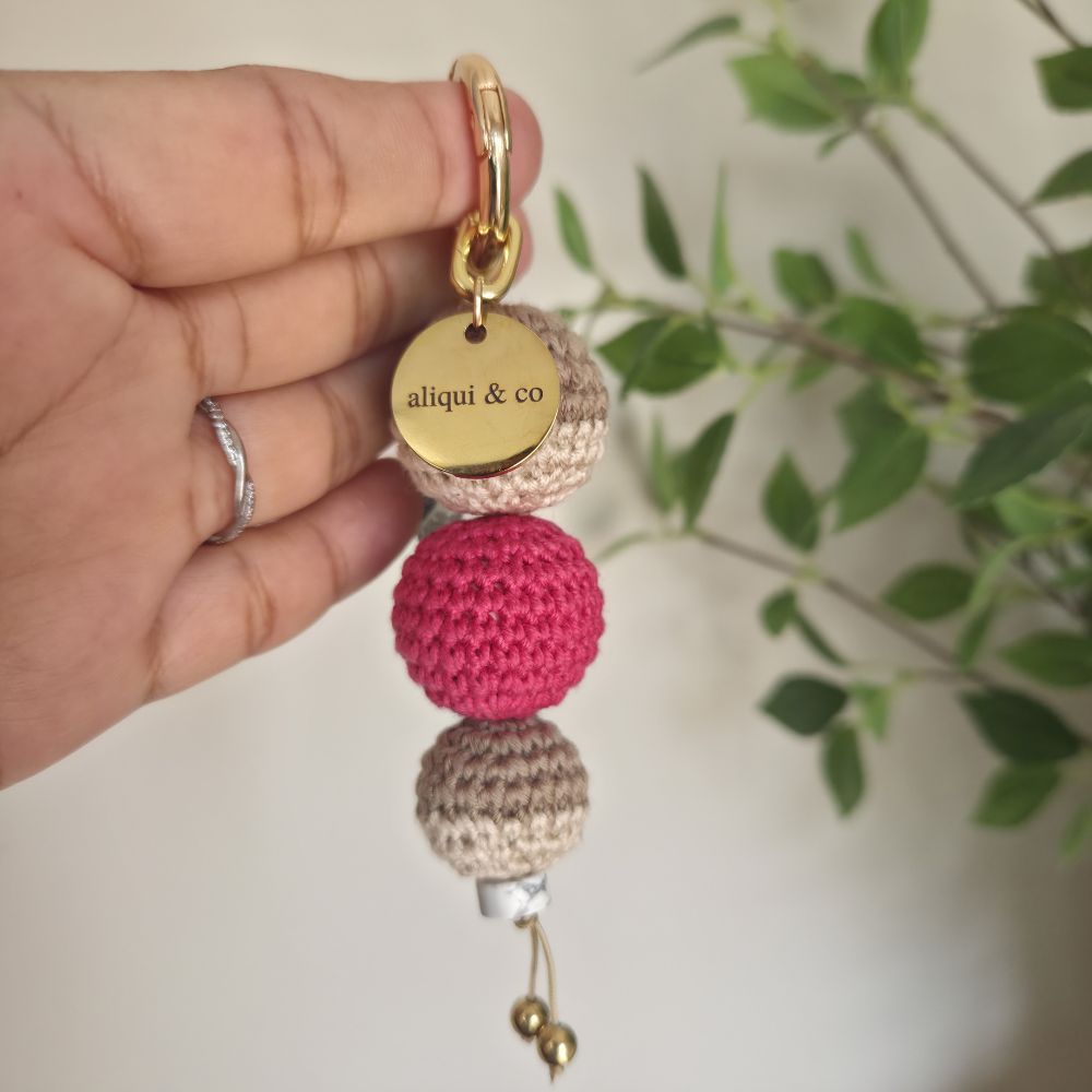 Hand holding a keychain with crocheted beads and a gold tag labeled 'aliqui & co'.