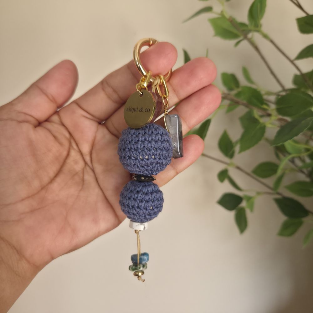 Hand holding a blue crocheted keychain with decorative elements against a blurred natural background