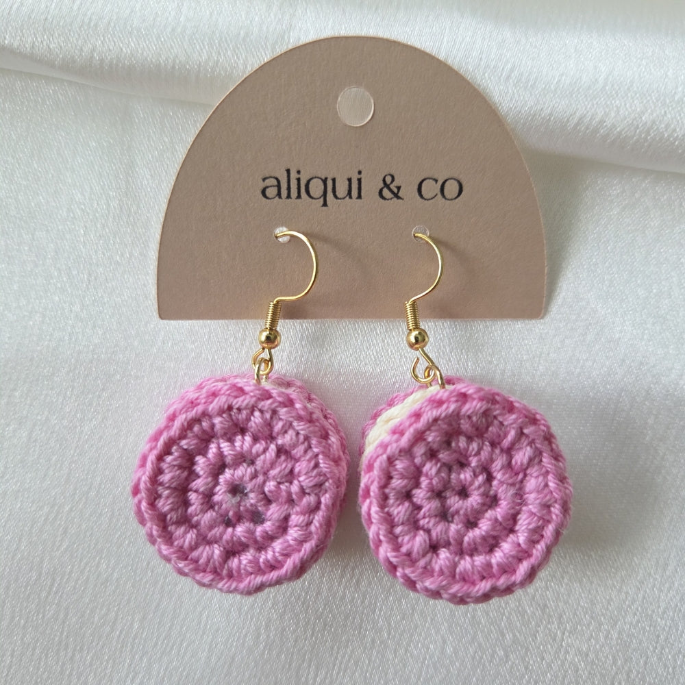 A pair of pink crocheted round drop earrings with gold fishhook wires, displayed on a brown paper hanger card printed with the brand name 'aliqui & co' against a white fabric background.