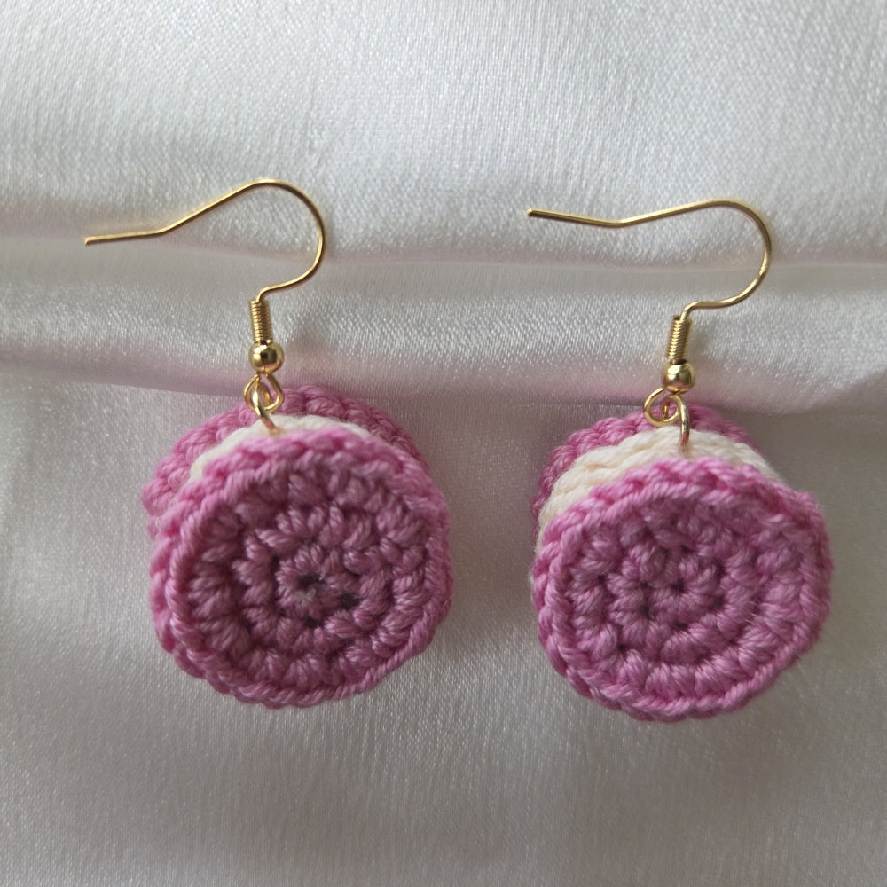 A pair of handmade crochet drop earrings shaped like pink and white sandwich cookies, hanging from gold fishhook wires on a white fabric background.