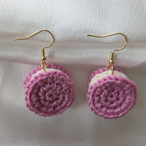 A pair of handmade crochet drop earrings shaped like pink and white sandwich cookies, hanging from gold fishhook wires on a white fabric background.