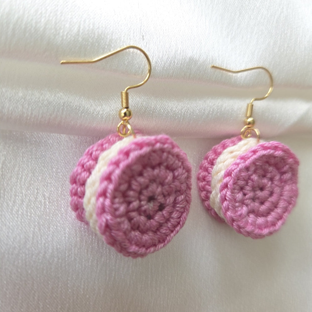A pair of handmade crochet dangle earrings shaped like pink and white macarons, suspended from gold fishhook wires, displayed on a soft white fabric background.