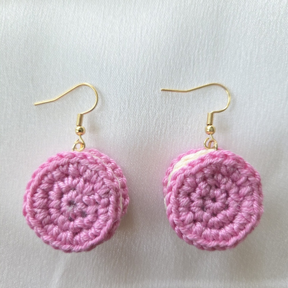A pair of handmade pink crochet dangle earrings with gold fishhook wires, displayed on a textured white fabric background.