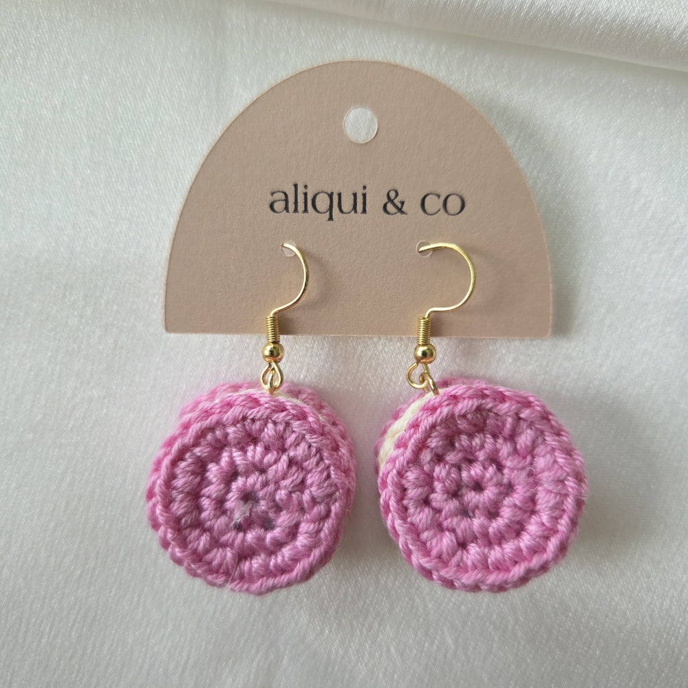 A pair of pink crocheted dangle earrings with gold fishhook wires, displayed on a beige paper hanger card printed with the brand name 'aliqui & co'. The earrings are photographed against a soft, textured white fabric background.