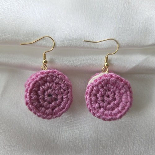 A pair of handmade pink crocheted dangle earrings with gold fishhook wires, displayed on a soft white fabric background.