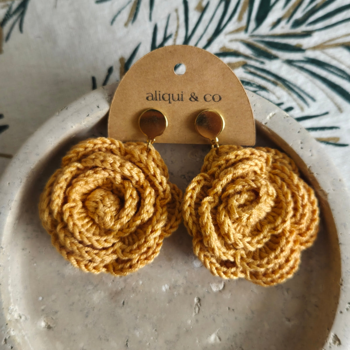 Pair of yellow crochet earrings on a 'aliqui & co' branded card, displayed on a textured surface with a leafy background.
