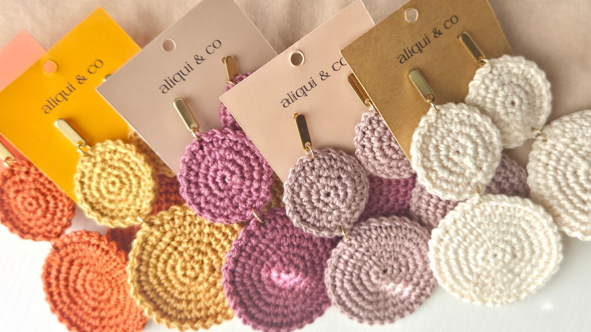 A set of handmade crocheted disc earrings in multiple colors, including yellow, orange, pink, purple, and cream, each attached to a paper display card with the brand name 'aliqui & co'.