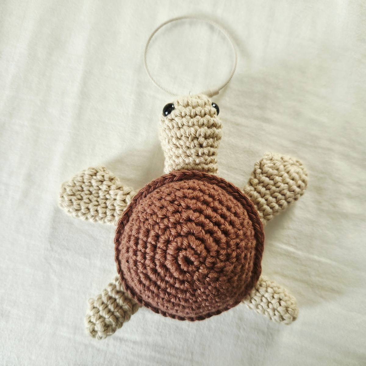 A handmade crocheted turtle toy with a brown spiral shell and cream-colored legs, head, and flippers, attached to a thin white hanging loop, laid flat on a white fabric background.
