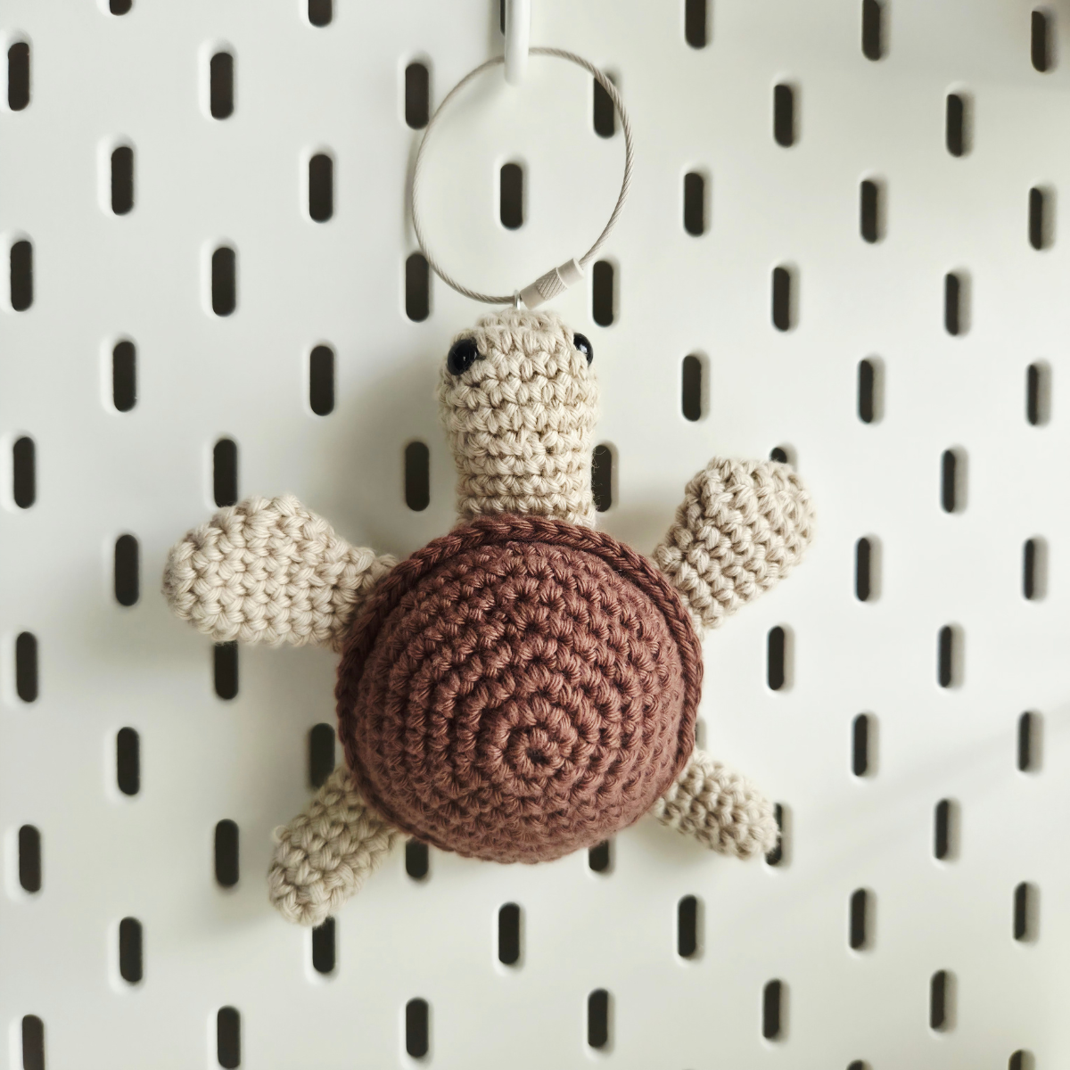 A crocheted turtle ornament with a brown shell and cream-colored limbs and head, hanging from a metal ring on a white pegboard background.