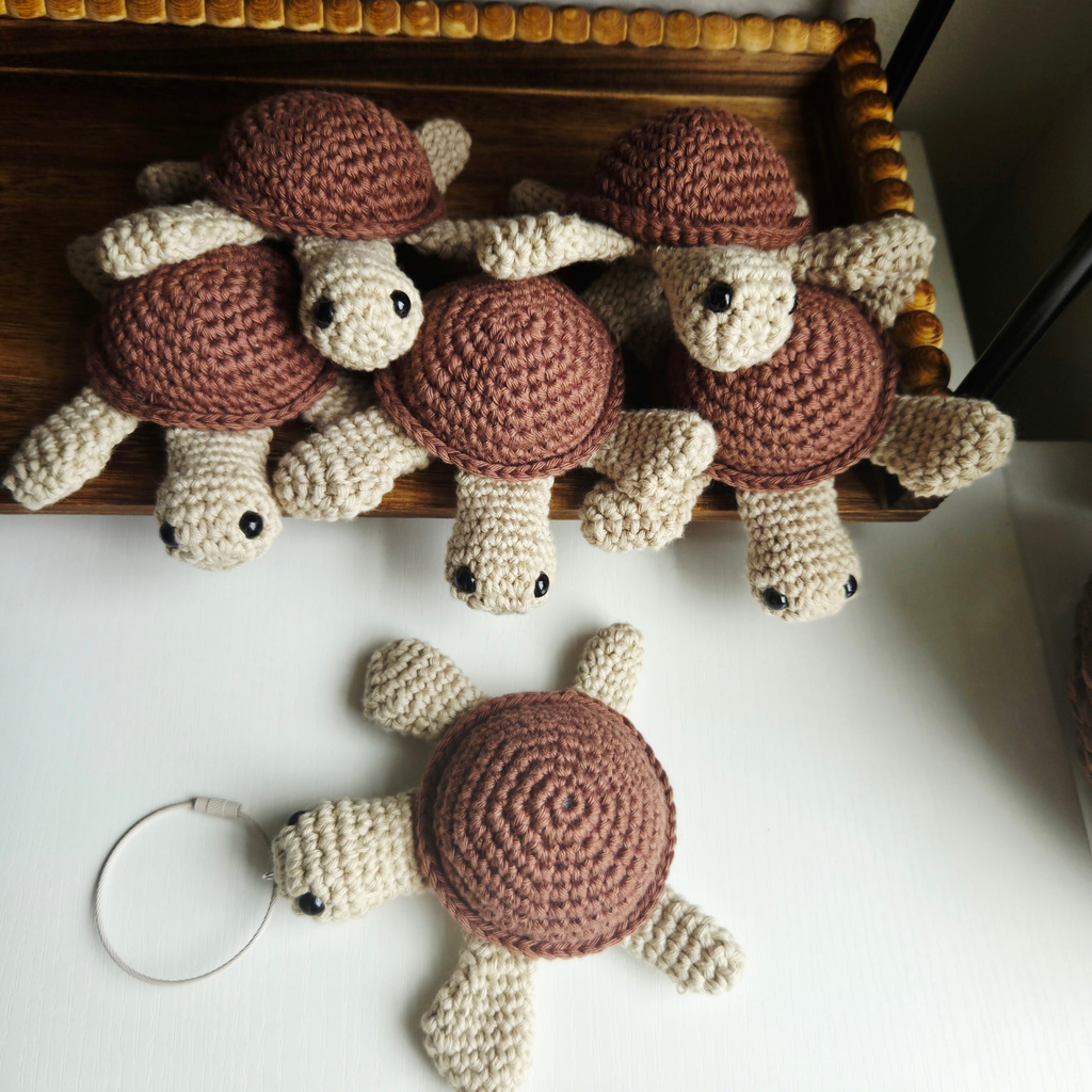 A set of handmade crocheted turtle keychains in brown and cream, displayed on a wooden tray and one lying on a white surface below.