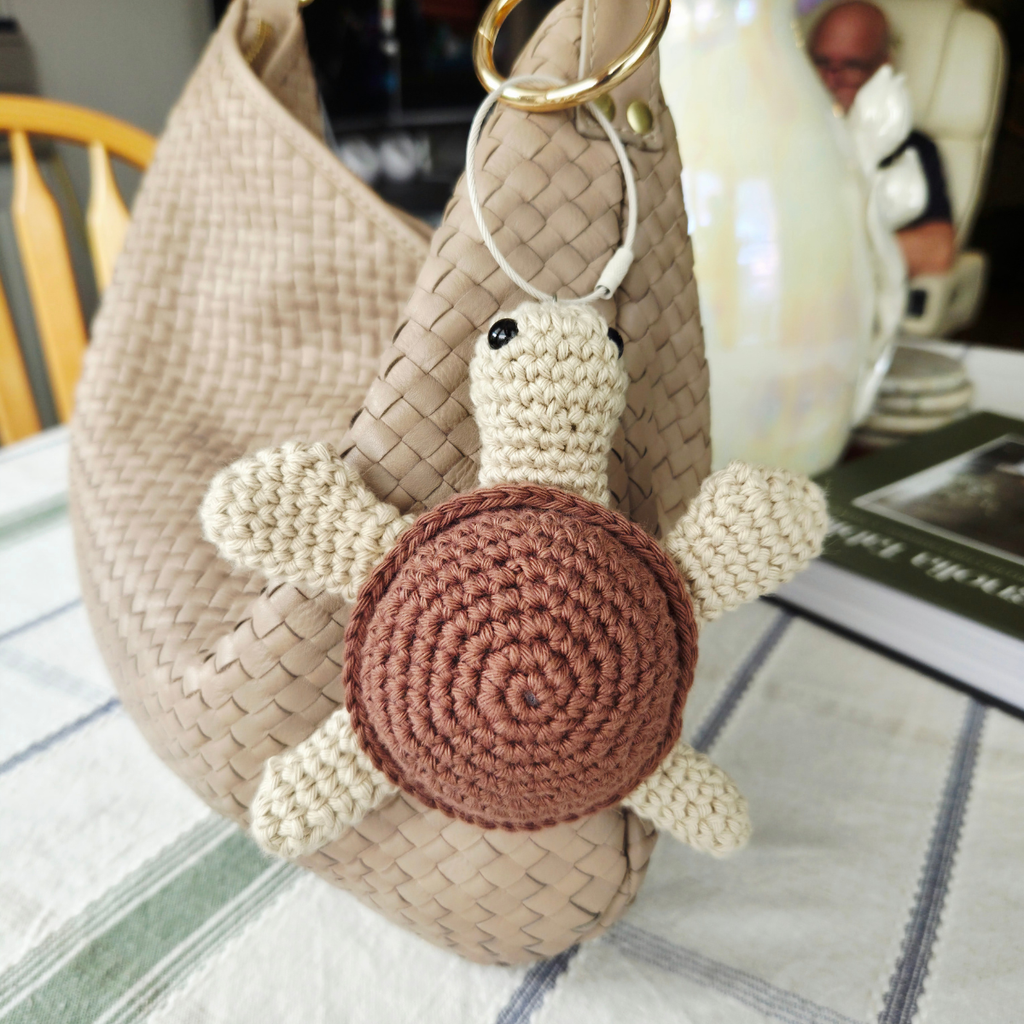A crocheted turtle keychain charm with a brown spiral shell and cream-colored limbs and head, attached to a woven beige shoulder bag with a gold metal ring, set on a table.