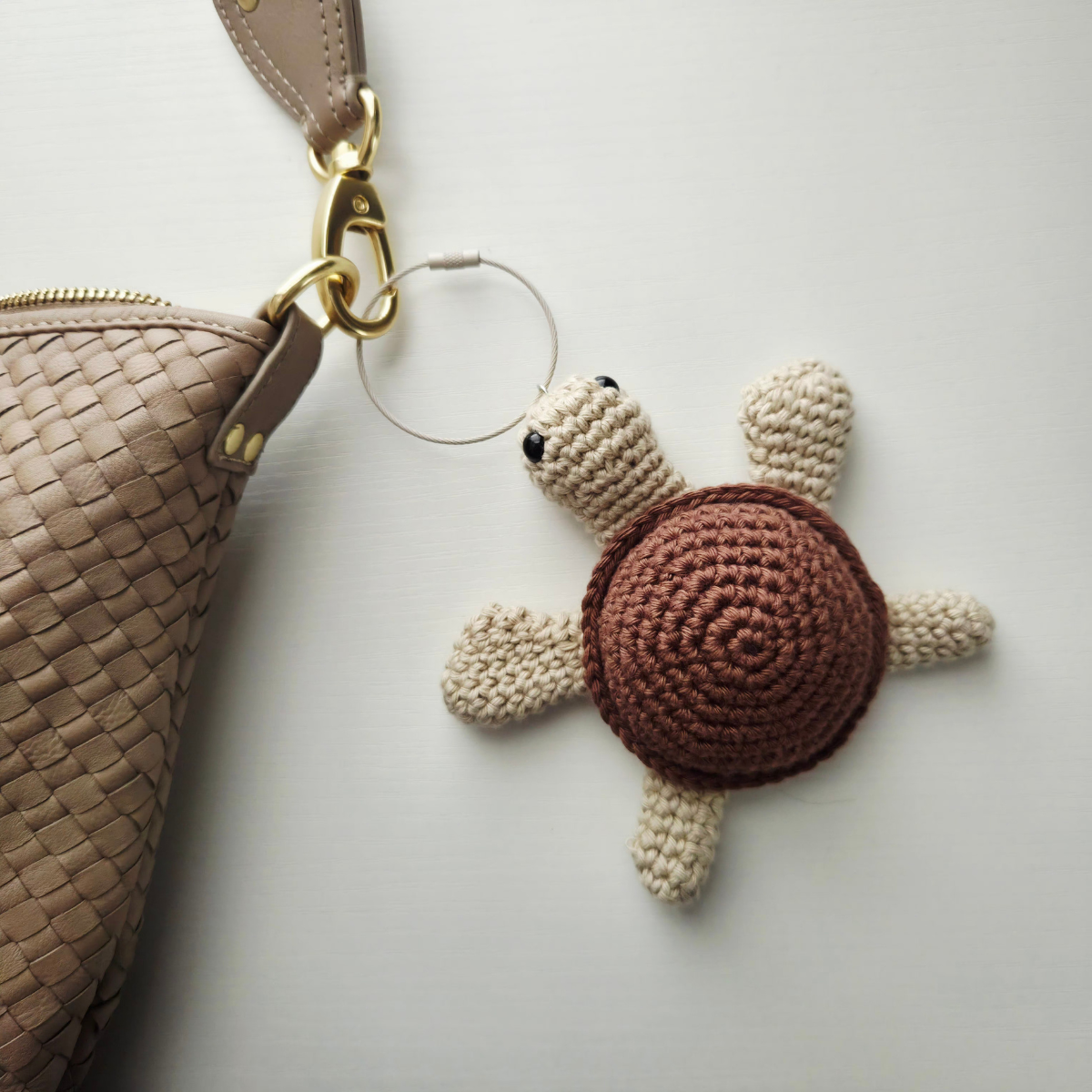 A crocheted sea turtle keychain charm with a brown shell and cream-colored flippers and head, attached to a clear key ring and shown next to a woven beige handbag on a light gray background.