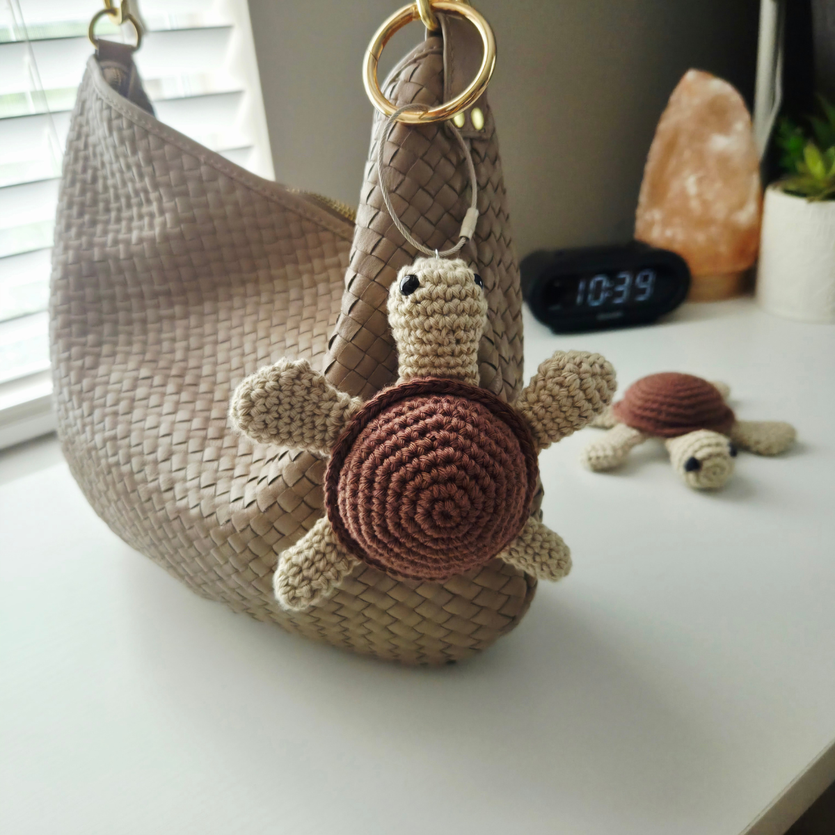A woven taupe shoulder bag with a crocheted turtle charm hanging from a gold ring, displayed on a white tabletop with a small matching crocheted turtle figurine in the background.