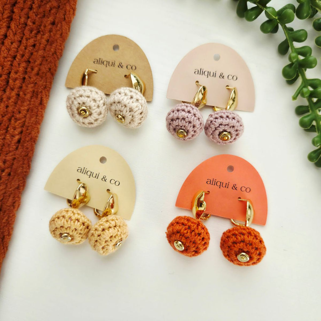 Four pairs of handmade crochet stud earrings displayed on arched cards. The earrings feature textured crochet balls in cream, beige, dusty mauve, and burnt orange, each with a small gold bead and gold-tone hoop.