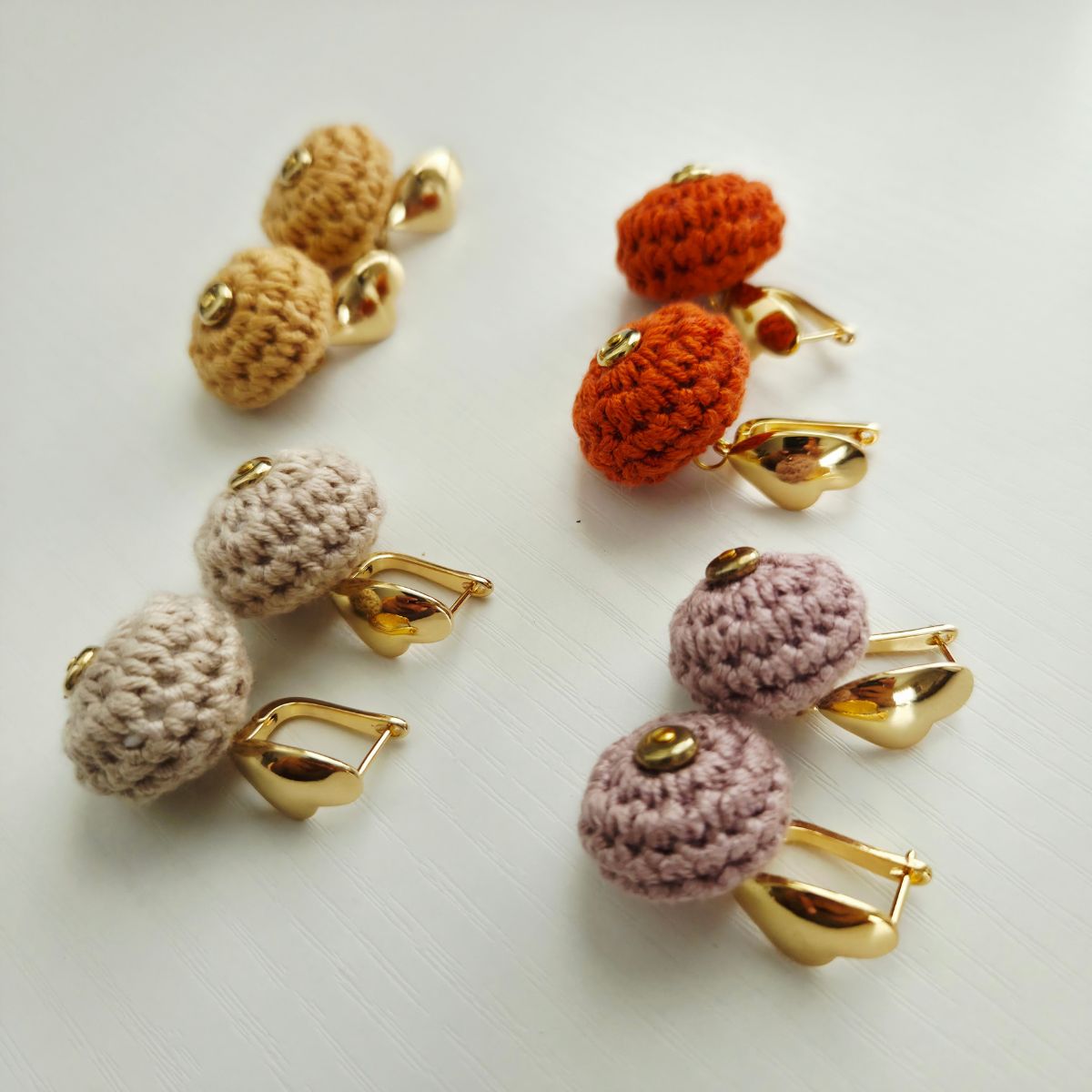 Four pairs of handmade crochet earrings with gold metal hooks, arranged on a light off-white background. The crochet tops are crocheted into ball shapes in beige, light taupe, orange, and dusty mauve, with small gold heart metal studs at the center.