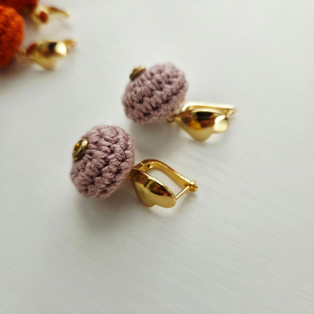 A pair of handmade crochet  ball earrings in a dusty mauve-pink color with gold metal hooks, displayed on a light gray textured surface.
