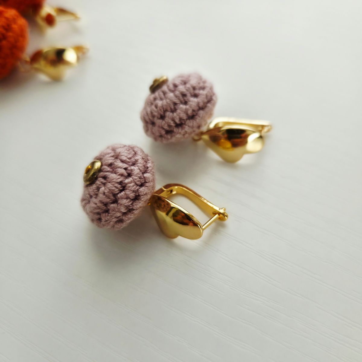 A pair of handmade crochet  ball earrings in a dusty mauve-pink color with gold metal hooks, displayed on a light gray textured surface.
