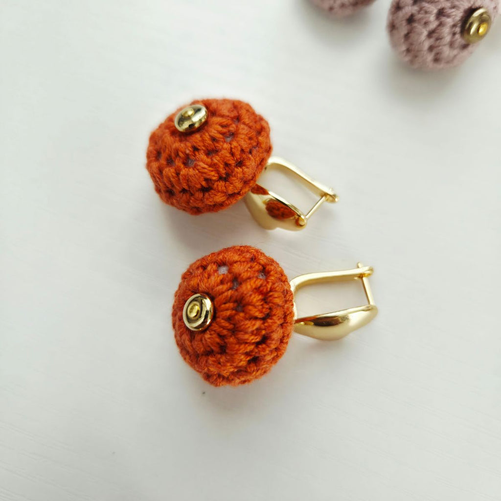 A pair of handmade orange crochet ball earrings with gold metal hooks and a small gold button, displayed on a light gray surface.