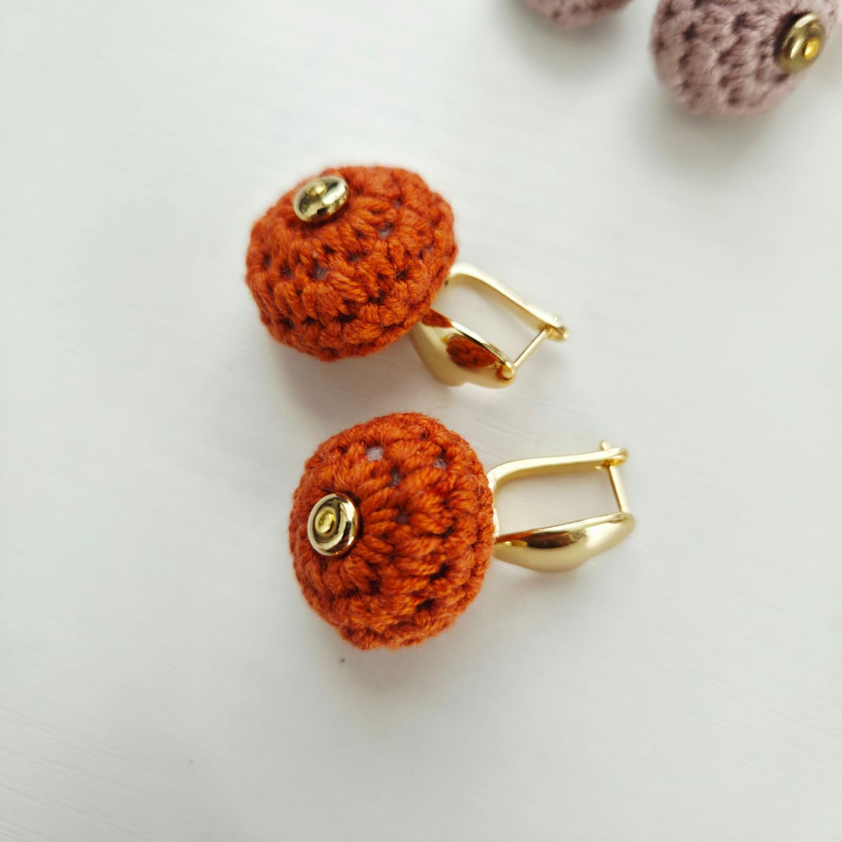 A pair of handmade orange crochet ball earrings with gold metal hooks and a small gold button, displayed on a light gray surface.