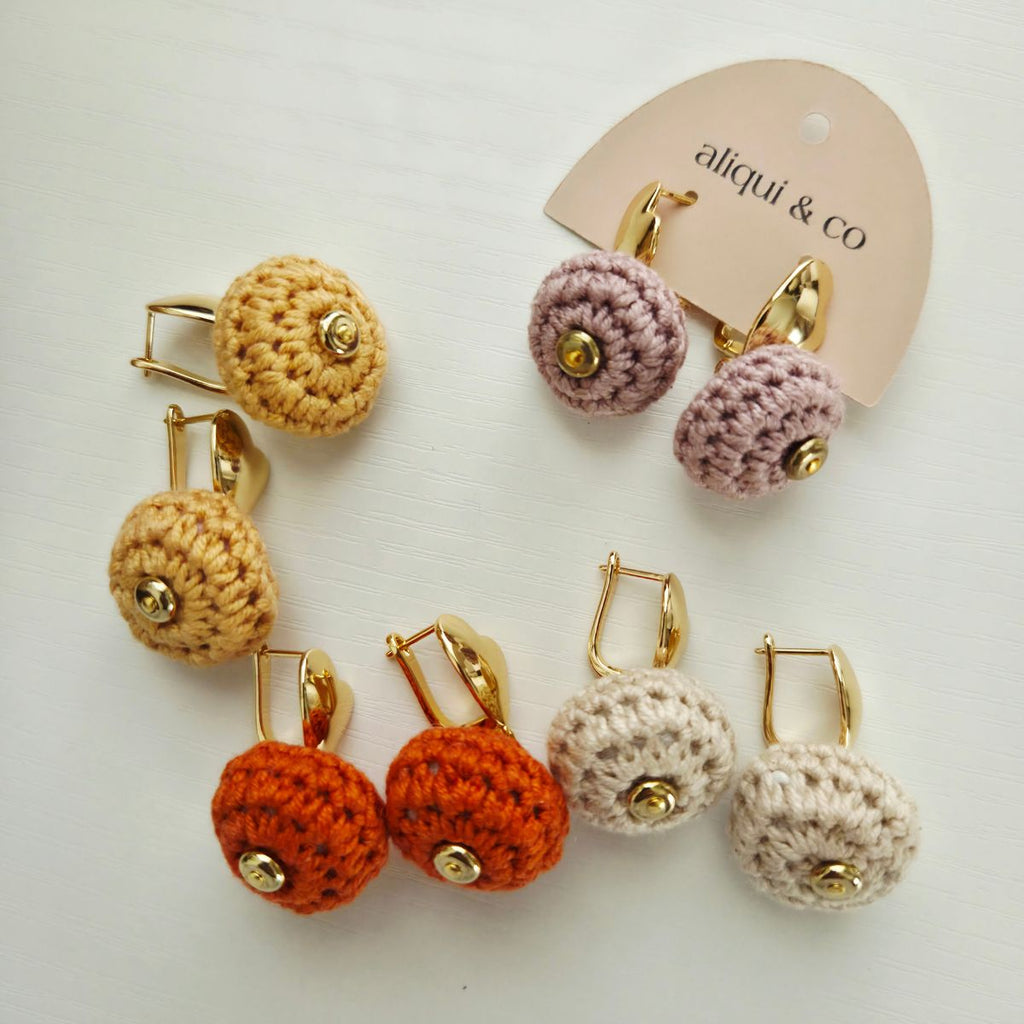 Four pairs of handmade crochet earrings with gold-tone metal hooks and round bead centers, displayed on a white background with a branded card that reads 'aliqui & co'. The earrings come in yellow, pink, orange, and cream colors.