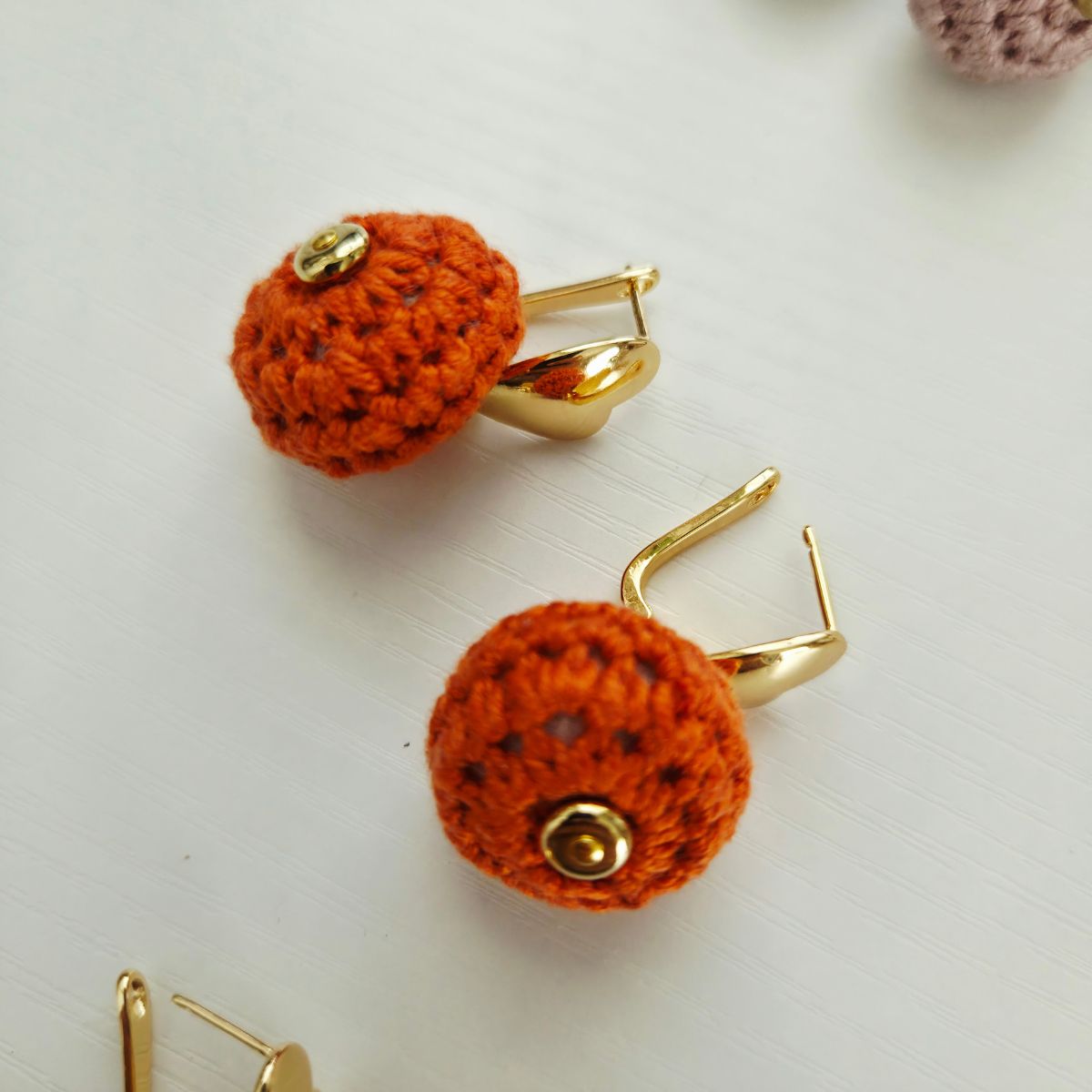 Orange crochet ball earrings with gold metal hooks and small gold beads, shown on a light gray surface.